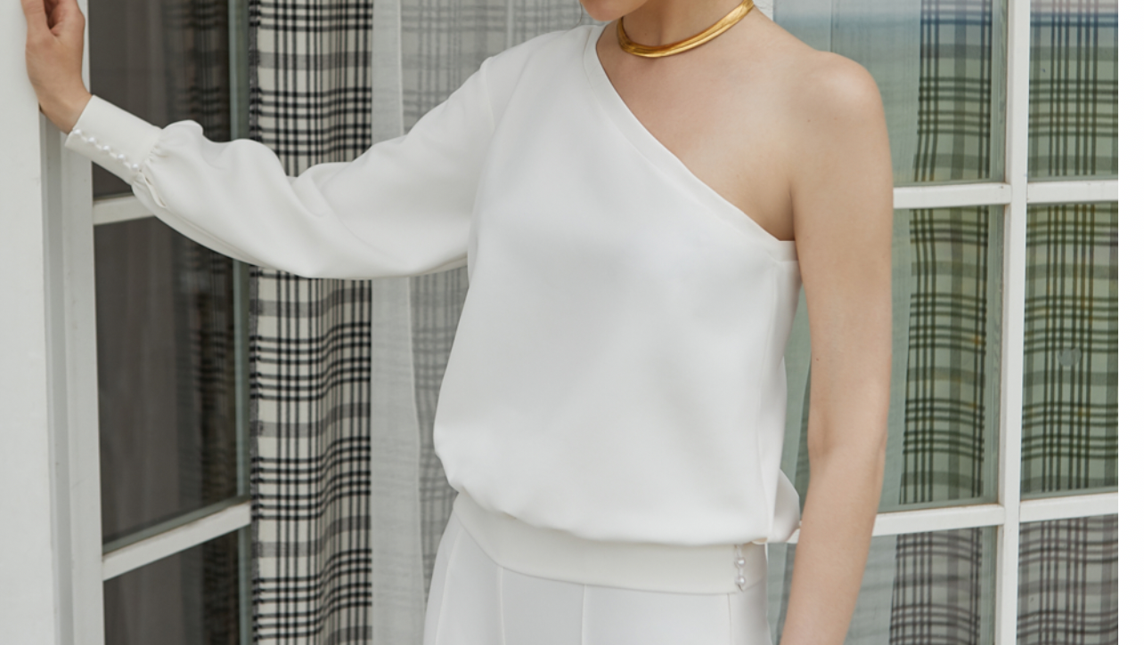 Eine Frau in einem weißen One-Shoulder-Top und einer weißen Hose steht vor einem Fenster.