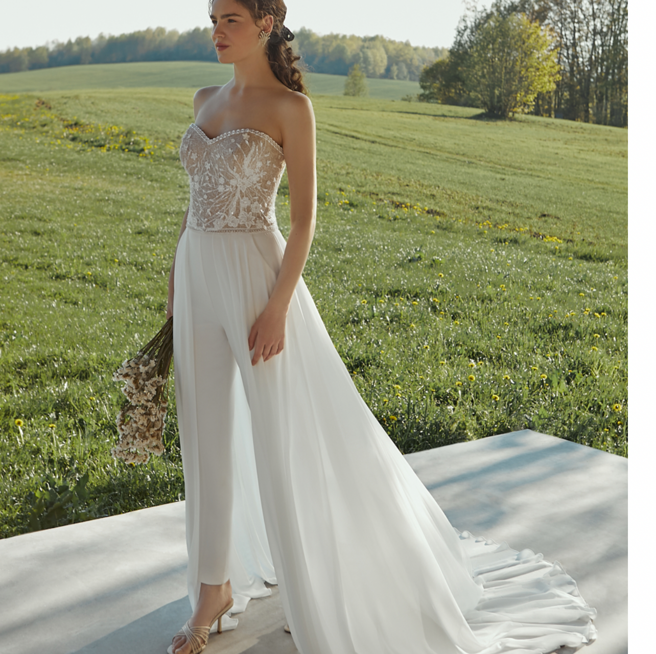 Eine Frau in einem weißen Brautkleid steht mit einem Blumenstrauß in der Hand auf einem Feld.
