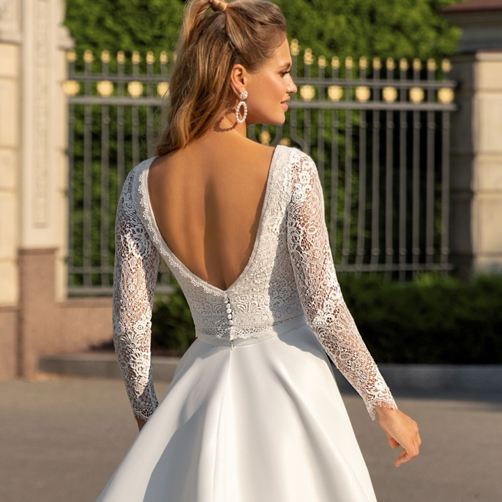 Eine Frau in einem weißen Brautkleid mit langen Ärmeln steht vor einem Zaun.