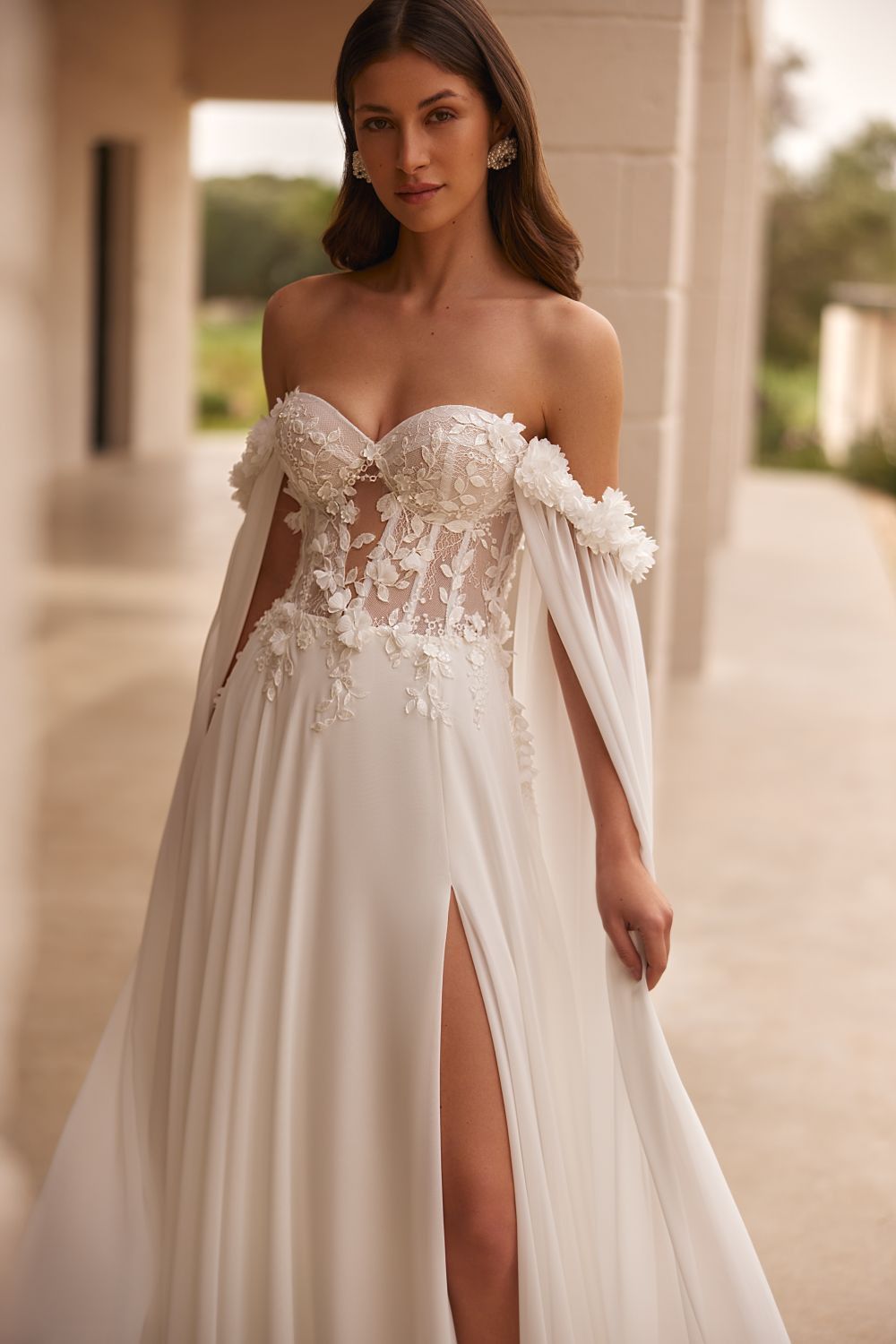 Eine Frau trägt ein weißes Brautkleid mit hohem Schlitz und einem Umhang.