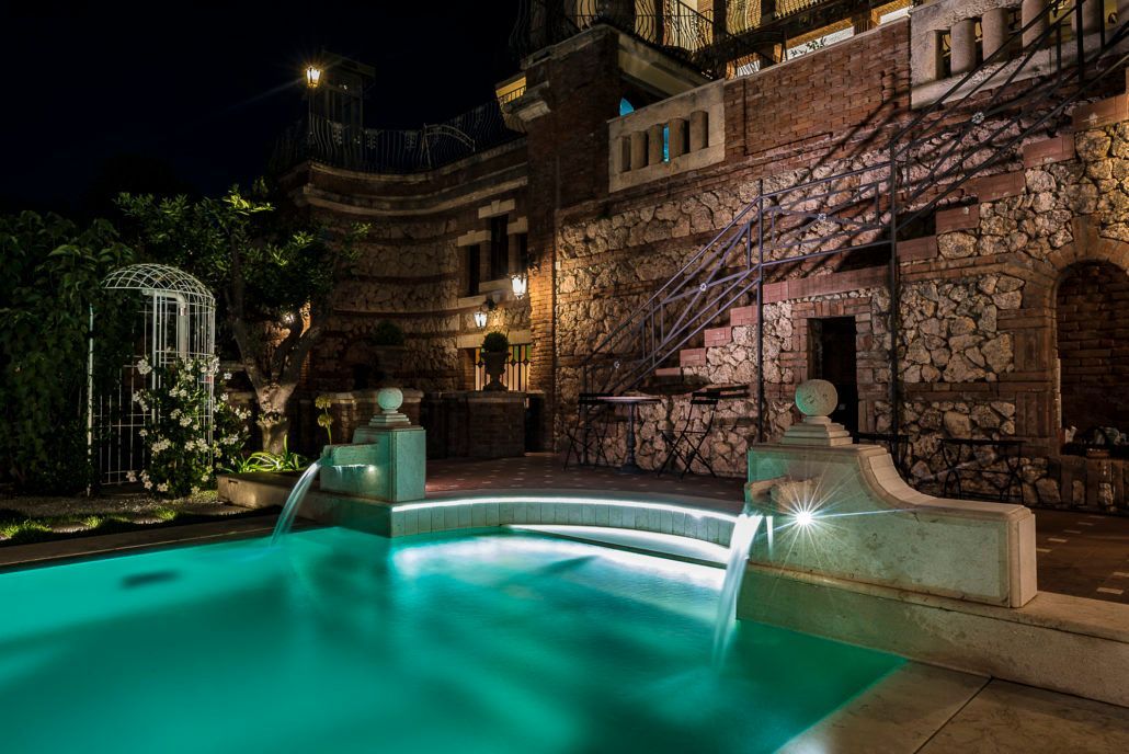 Una grande piscina è illuminata di notte di fronte a un edificio in pietra.