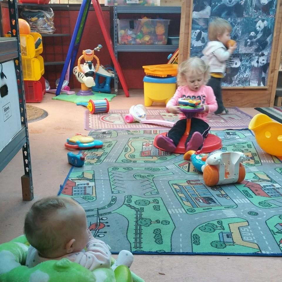 Children playing in a colorful playroom with toys, including a road-themed rug and an infant in a green chair.