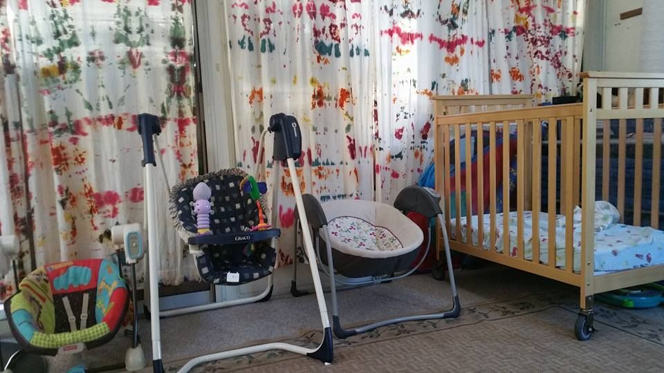 Baby equipment, including a swing, high chair, and crib, is set up in a room with patterned curtains.