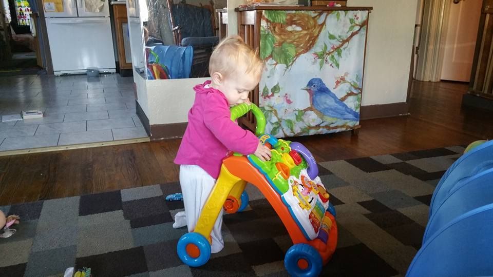 Blond toddler in pink top playing with a colorful baby walker on a patterned rug.