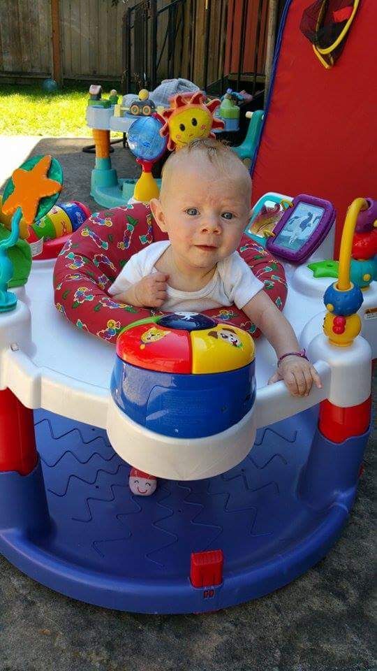 Baby in a colorful activity center outdoors, smiling, with toys and a fence in the background.