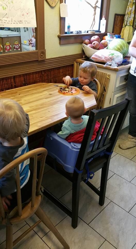 Three young children sit at a wooden table in a kitchen. One child uses a high chair.