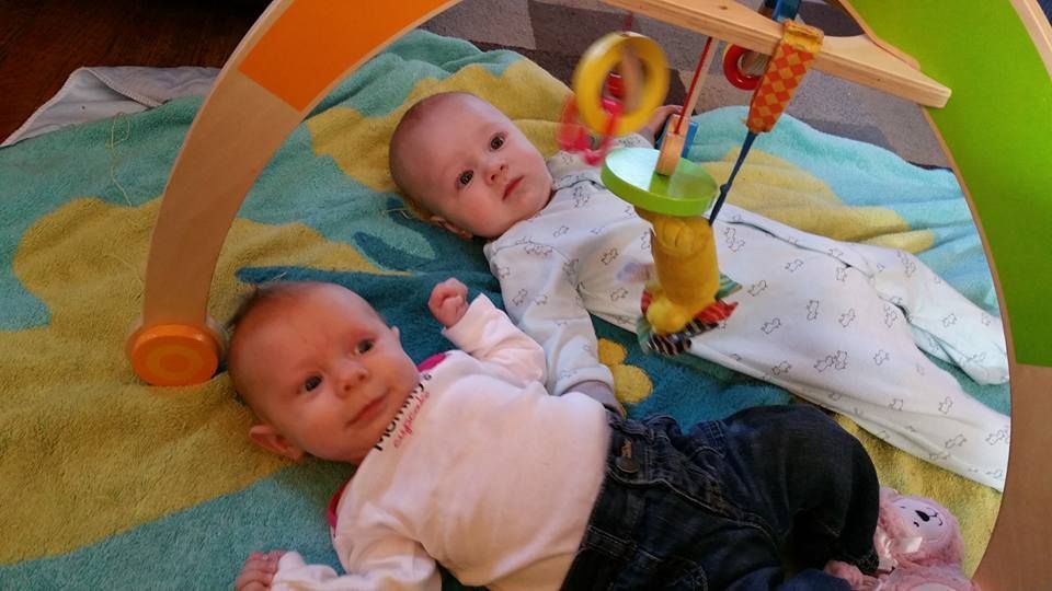 Two babies lying on a patterned mat under a wooden activity arch, looking at hanging toys.