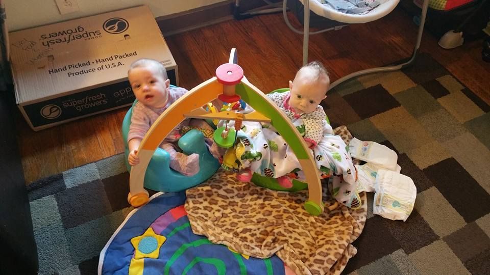 Two babies sitting near a play gym on a colorful mat; box in the background.