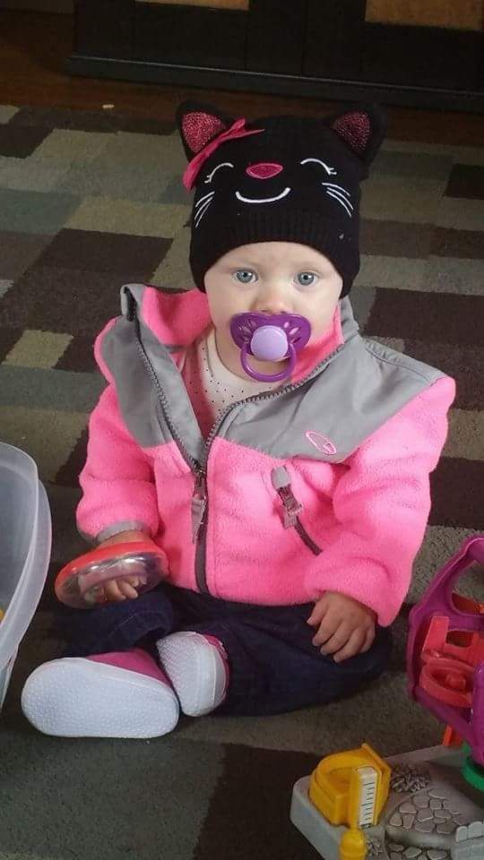 Child wearing a black cat beanie, pink jacket, and pacifier, sitting on a patterned rug.