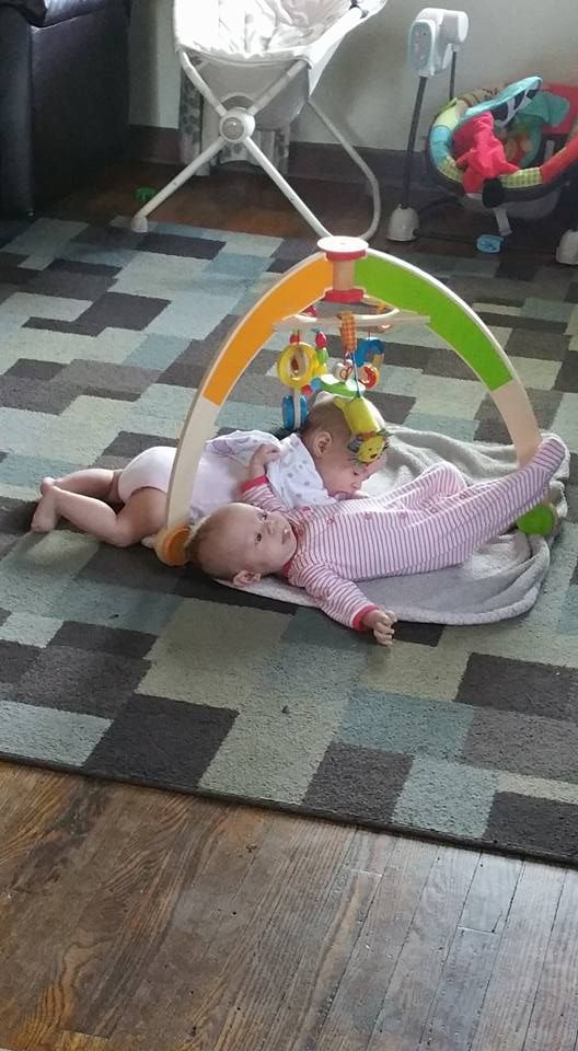 Two babies lying on a play mat under a toy arch on a patterned rug.