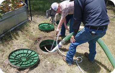 Two workers opening a green septic tank cover and hose on a grassy yard