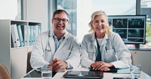 Two doctors in white coats smiling at a desk in a modern clinic, with medical charts on a monitor.