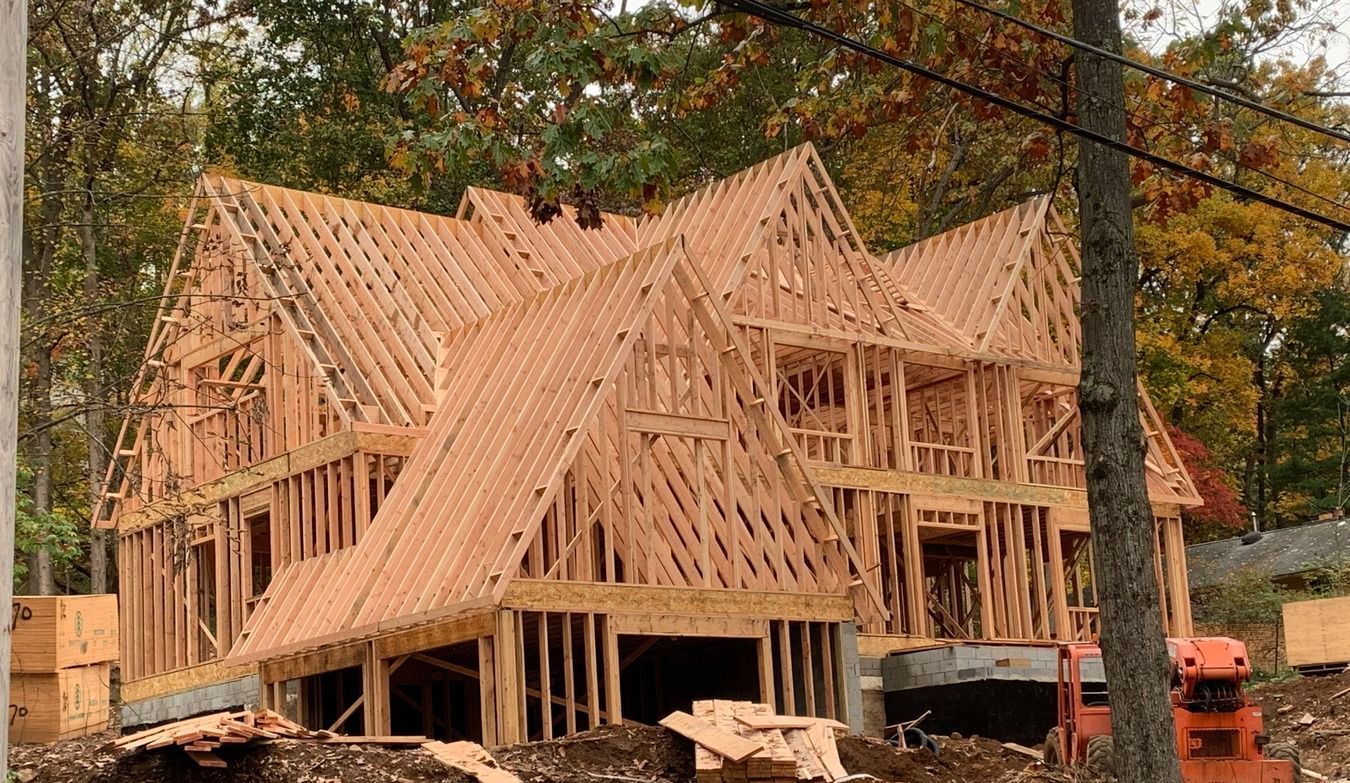 Wooden house frame under construction, surrounded by building materials, trees in background.