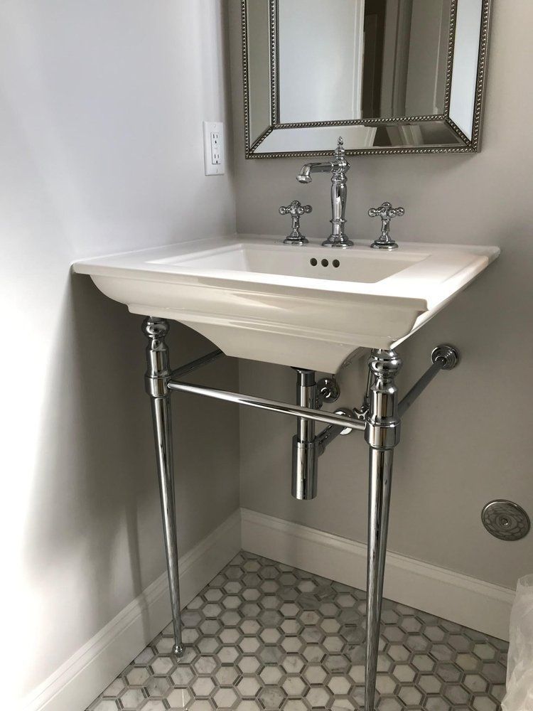 White pedestal sink with chrome legs and faucet, in a bathroom with hexagon tile floor.