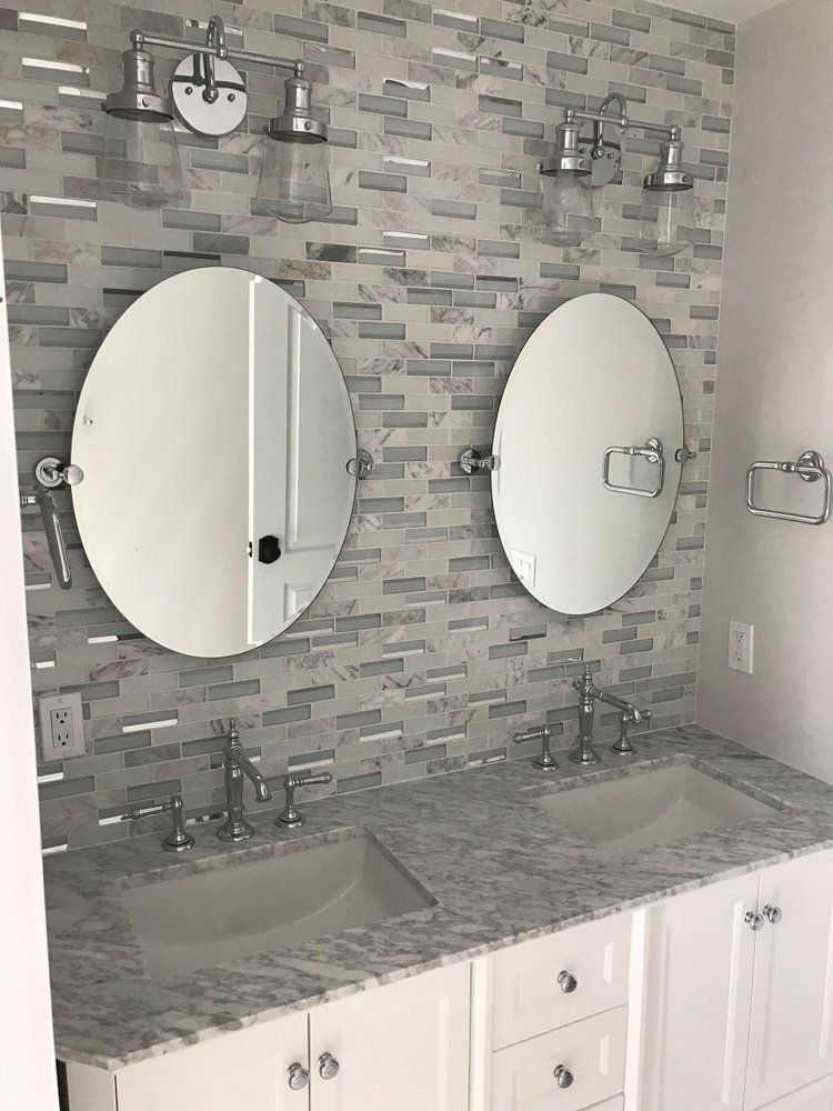 Bathroom with two oval mirrors, silver fixtures, white cabinets, and patterned tile backsplash.