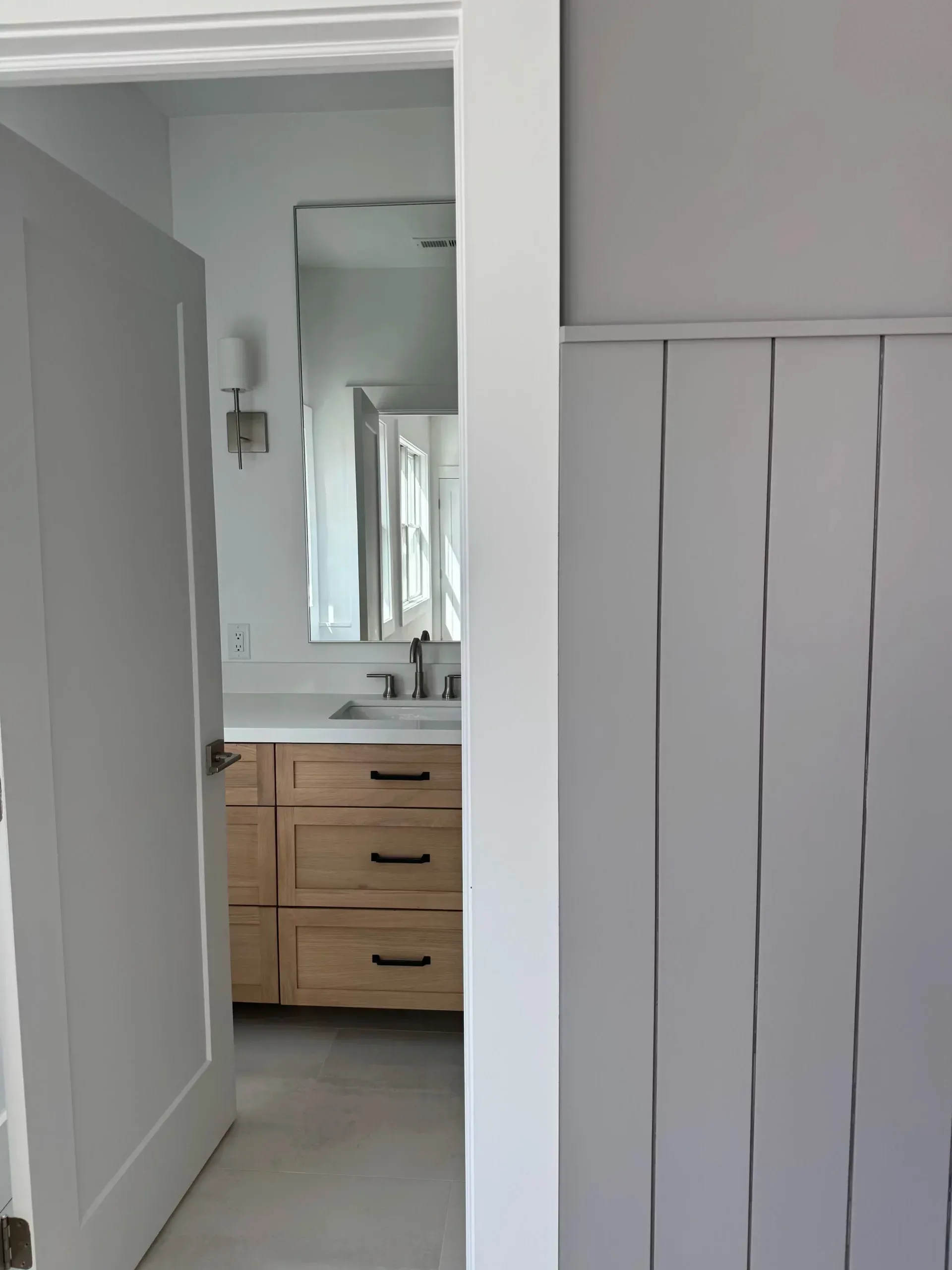 Bathroom interior with wood vanity, large mirror, and white wall with vertical paneling, viewed through doorway.