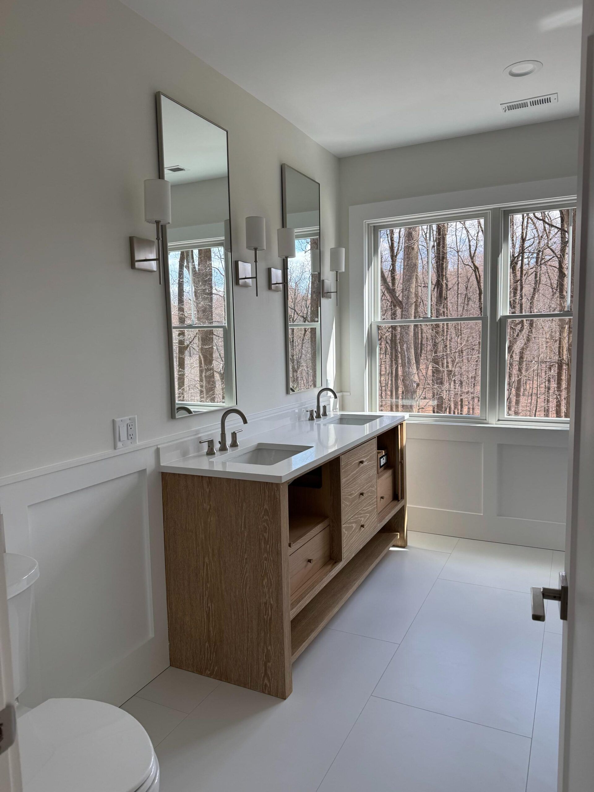 Bathroom with white walls, wood vanity, two sinks, mirrors, and large window overlooking trees.
