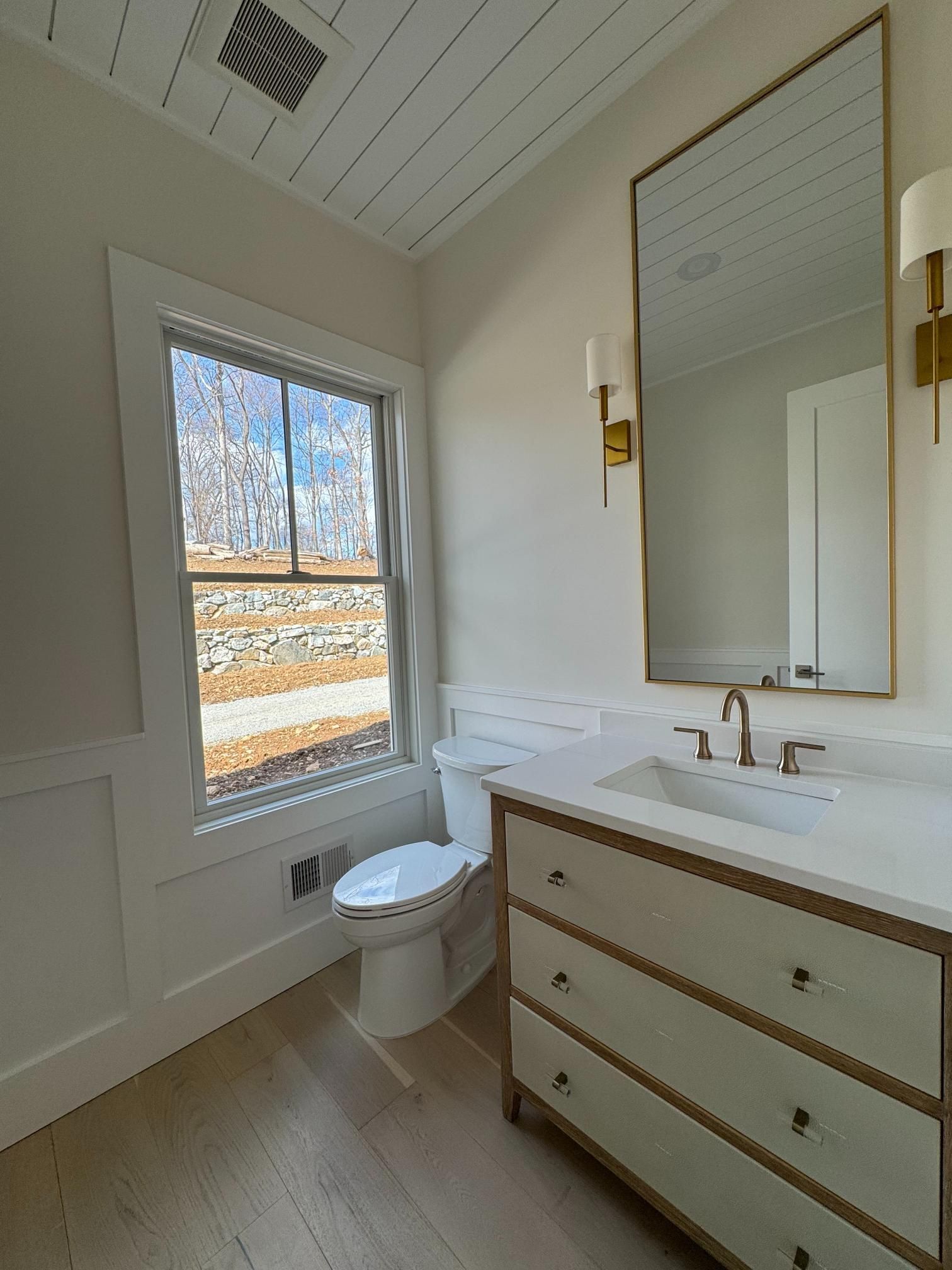 Bathroom with window, toilet, vanity, and large mirror; white, gold, and light wood tones.