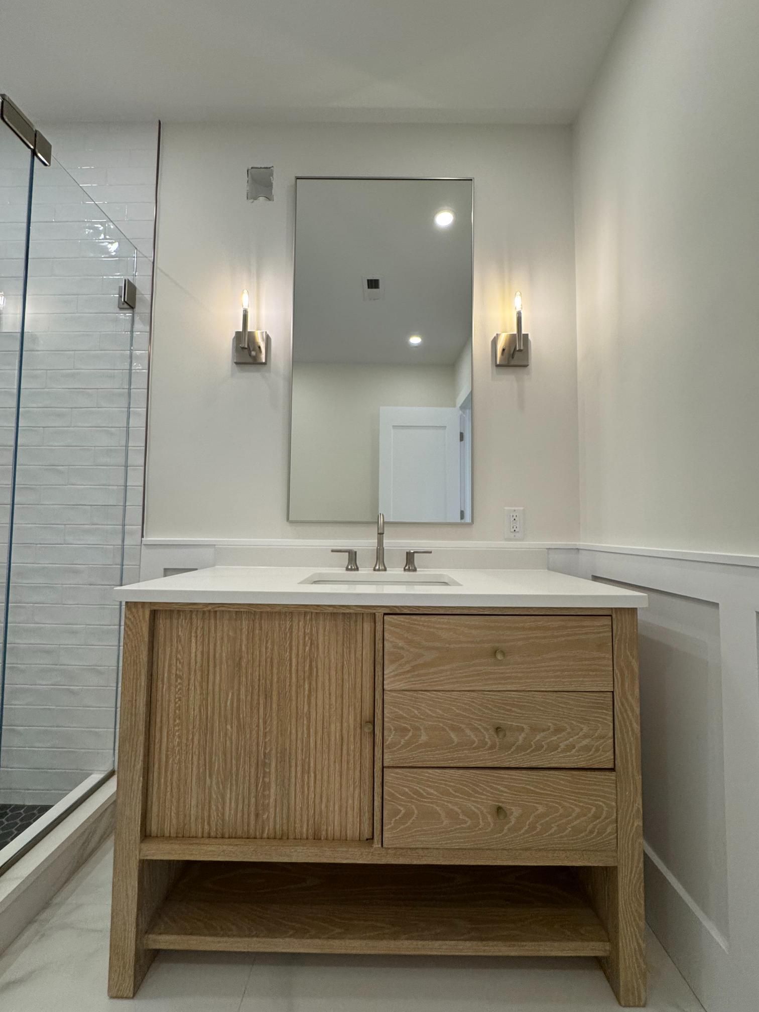 Wooden vanity with white countertop and mirror; sconces on white wall; shower to the left.
