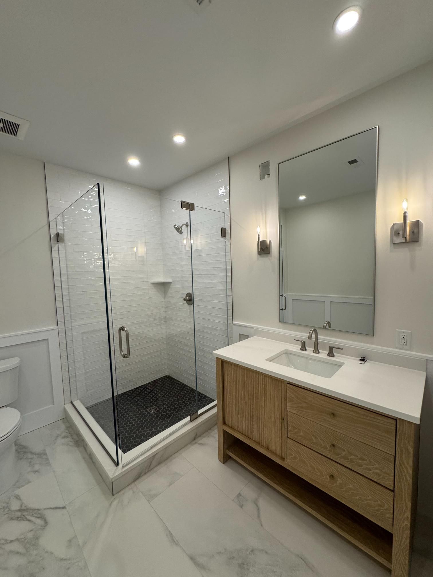 Modern bathroom with white walls, glass shower, wooden vanity, marble floor.