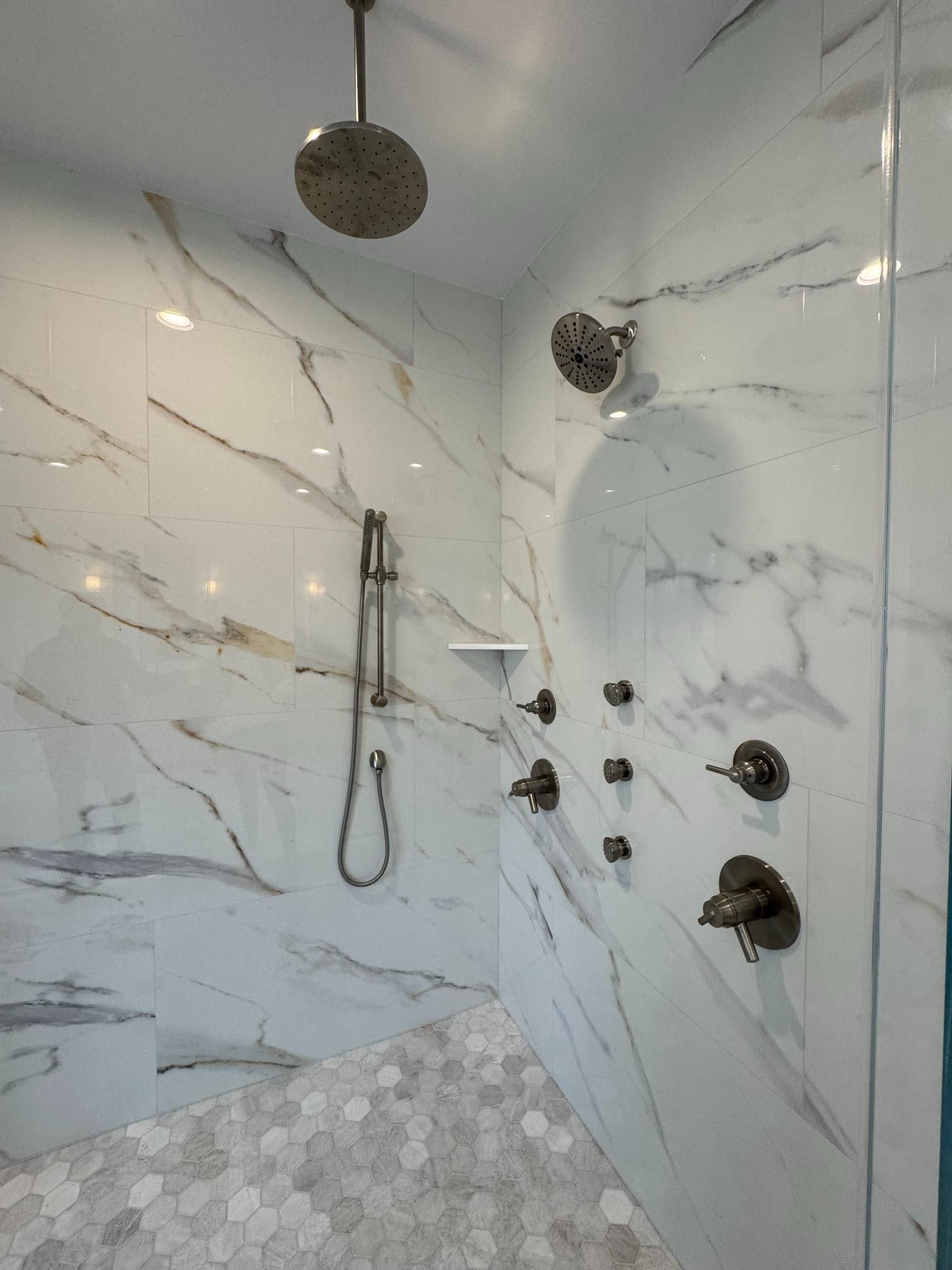 Luxury shower with marble-look tile, multiple showerheads, handheld shower, and hexagon tile floor.