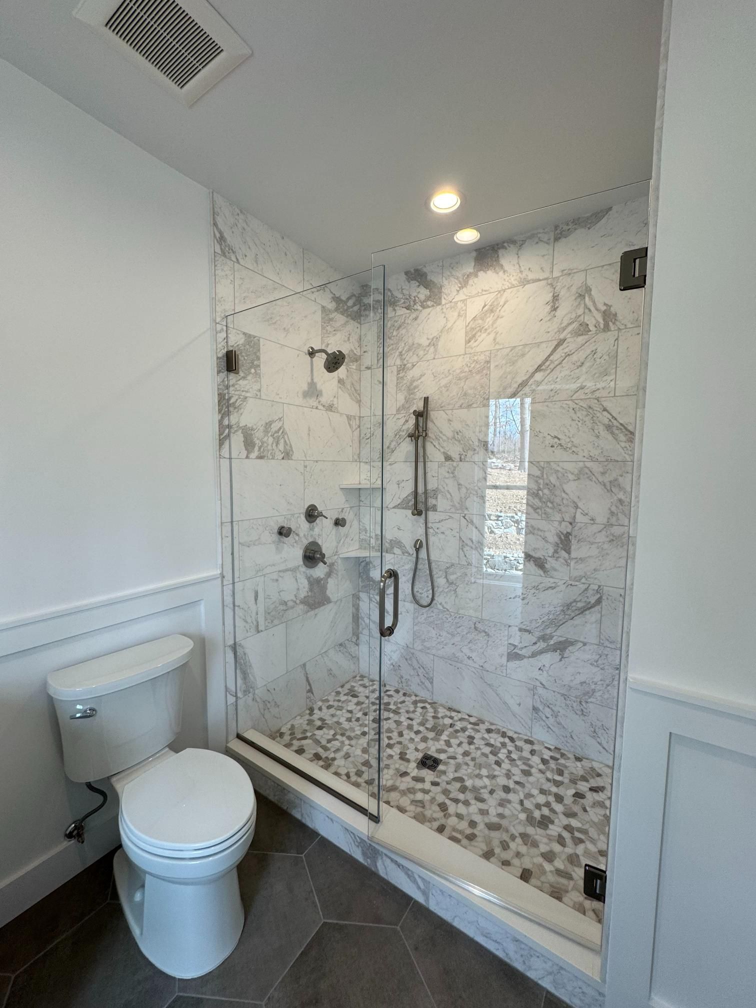 Bathroom with glass shower, marble tile, pebble floor, toilet. White walls and trim.