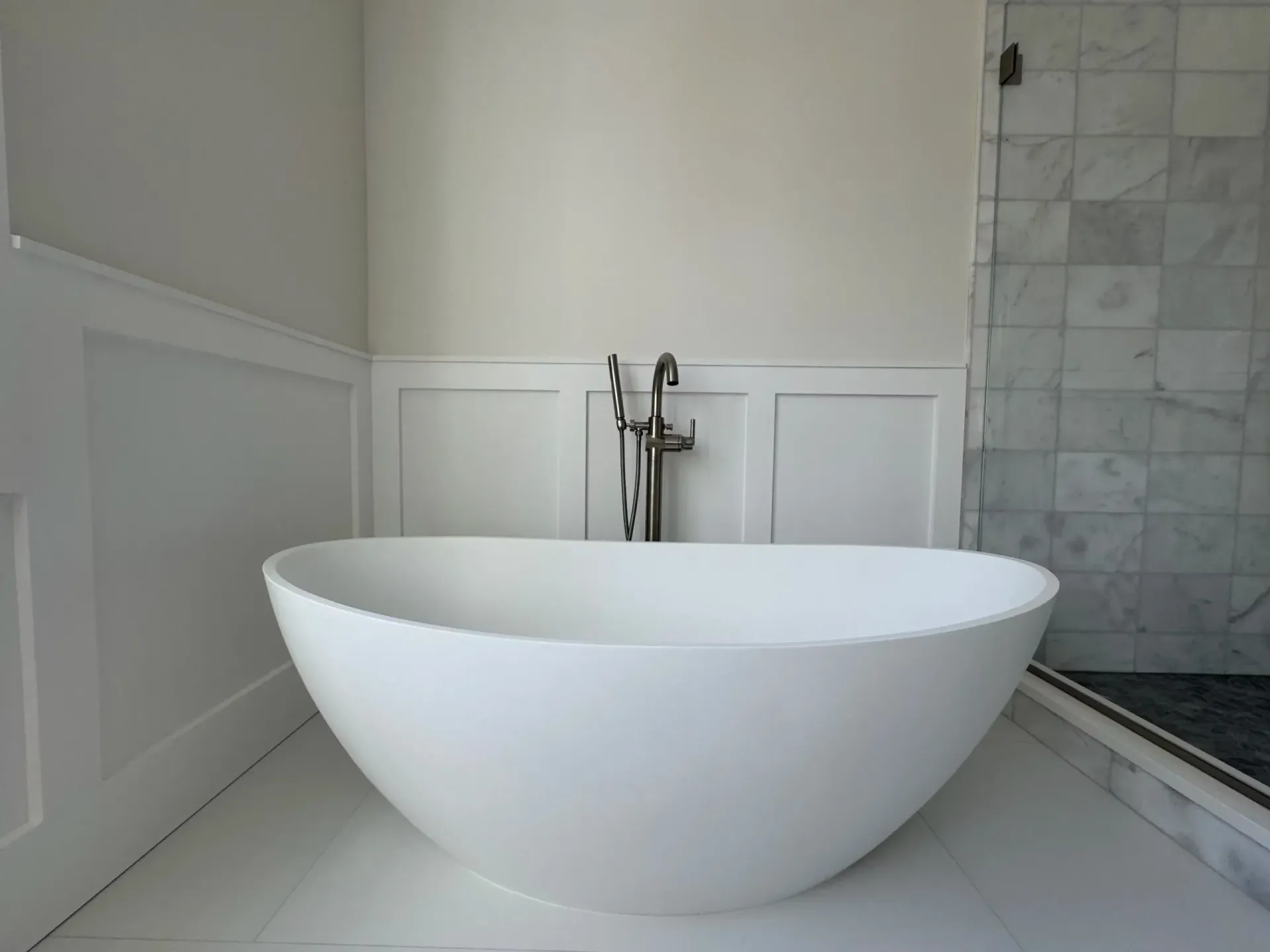 White oval bathtub in a bright bathroom with paneling and a marble shower.