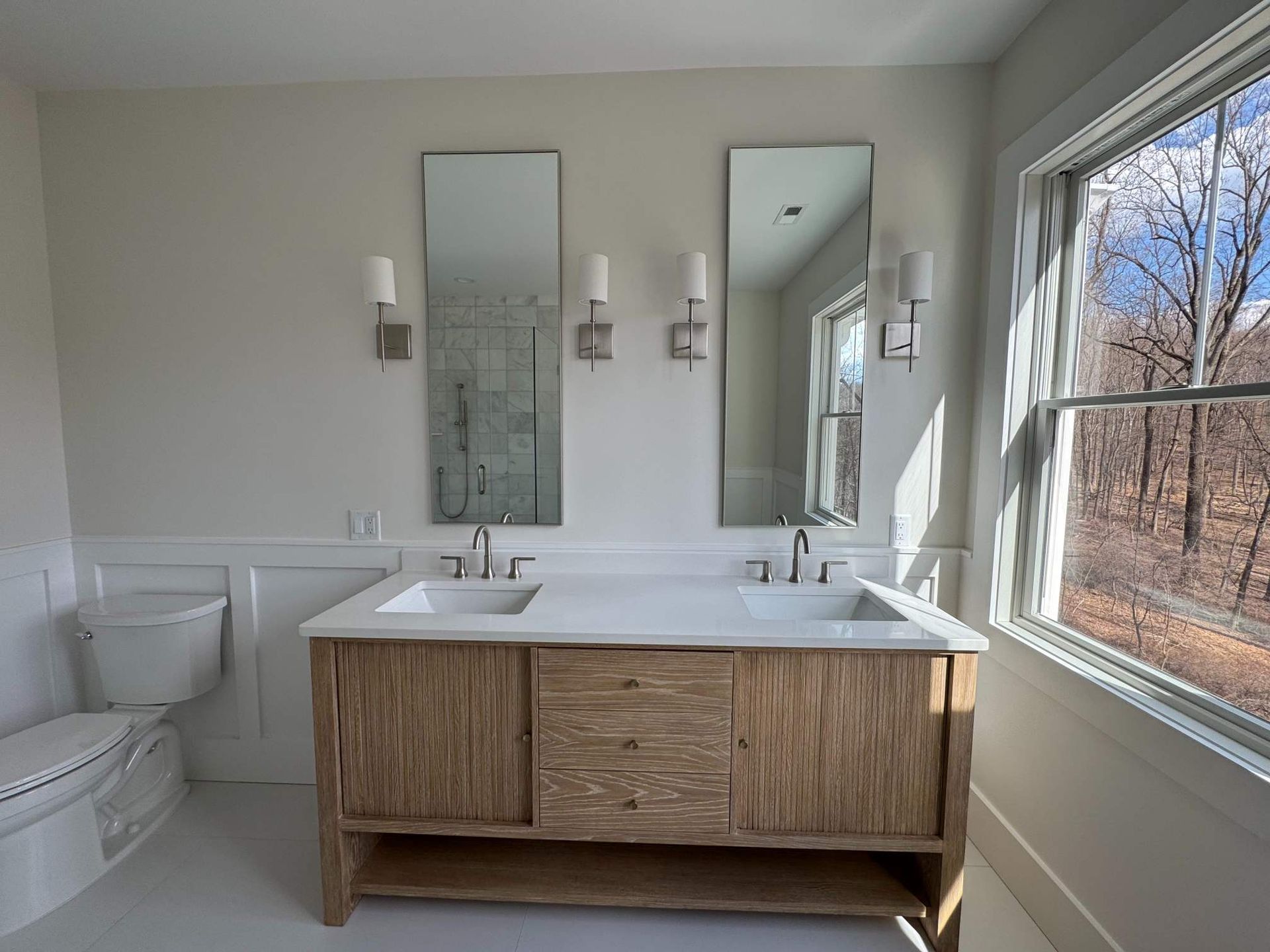 Bathroom with double vanity, two tall mirrors, and a window with a view of trees. Light wood and white accents.