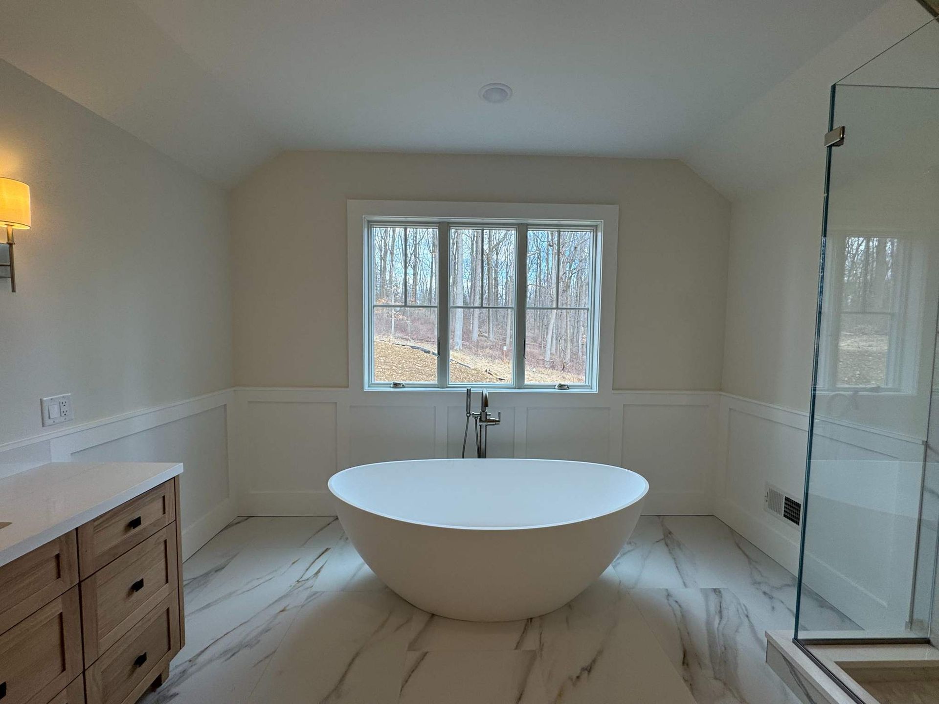 Spacious bathroom with a white oval tub, marble floor, and large window overlooking trees.