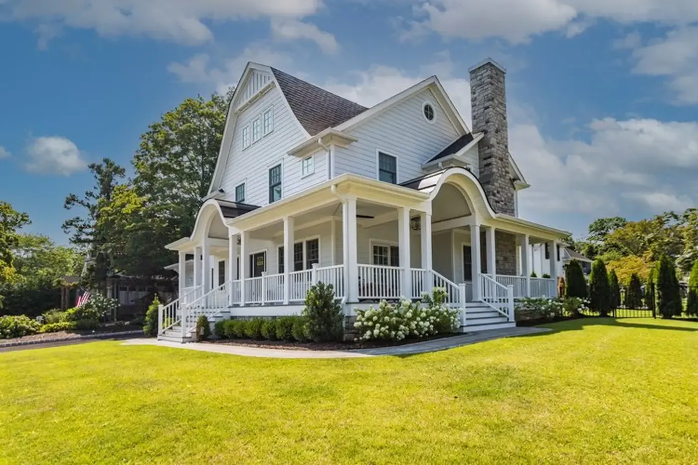 White farmhouse with wrap-around porch, stone chimney, and green lawn under a blue sky.