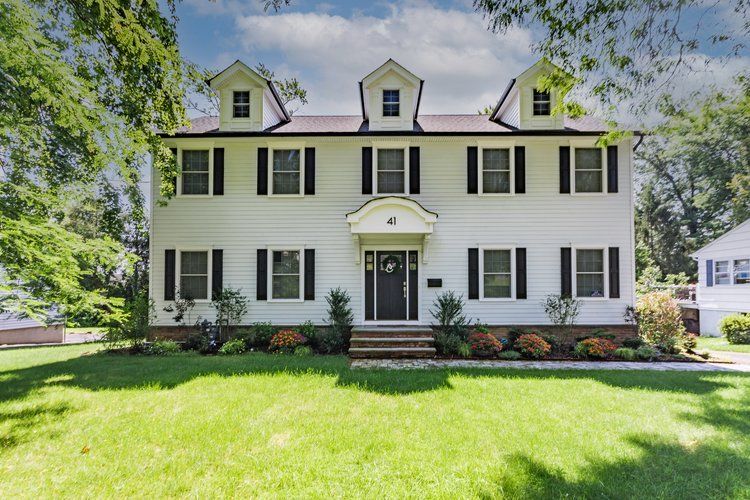 White two-story house with black shutters and a black door, green lawn, trees, and blue sky.