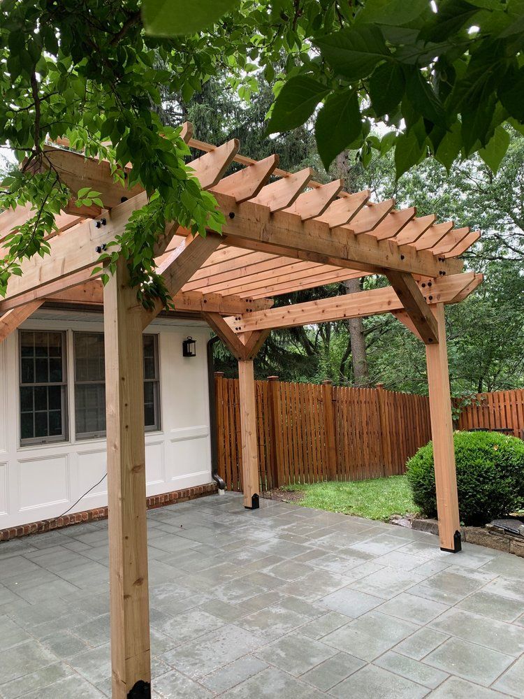 Wooden pergola over a patio. Light brown wood, surrounded by greenery and a brick building.