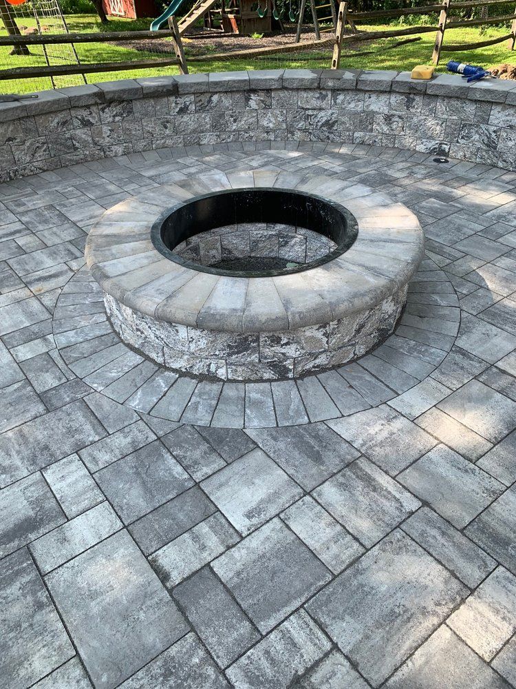 Stone patio with circular fire pit. Grey paving stones, dark metal ring, and stone retaining wall.
