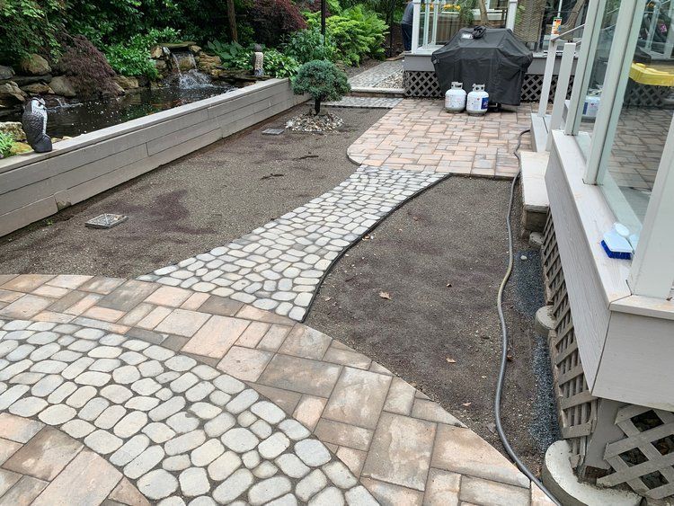 Stone paths lead through a yard with a pond, patio, and grill. Brown dirt and green plants are also present.