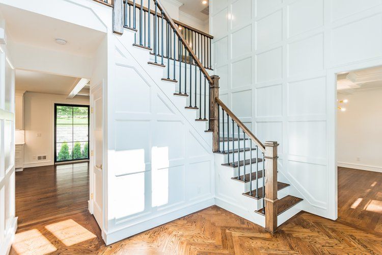 Wooden staircase with dark railing and white panel walls in a bright entryway with hardwood floors.