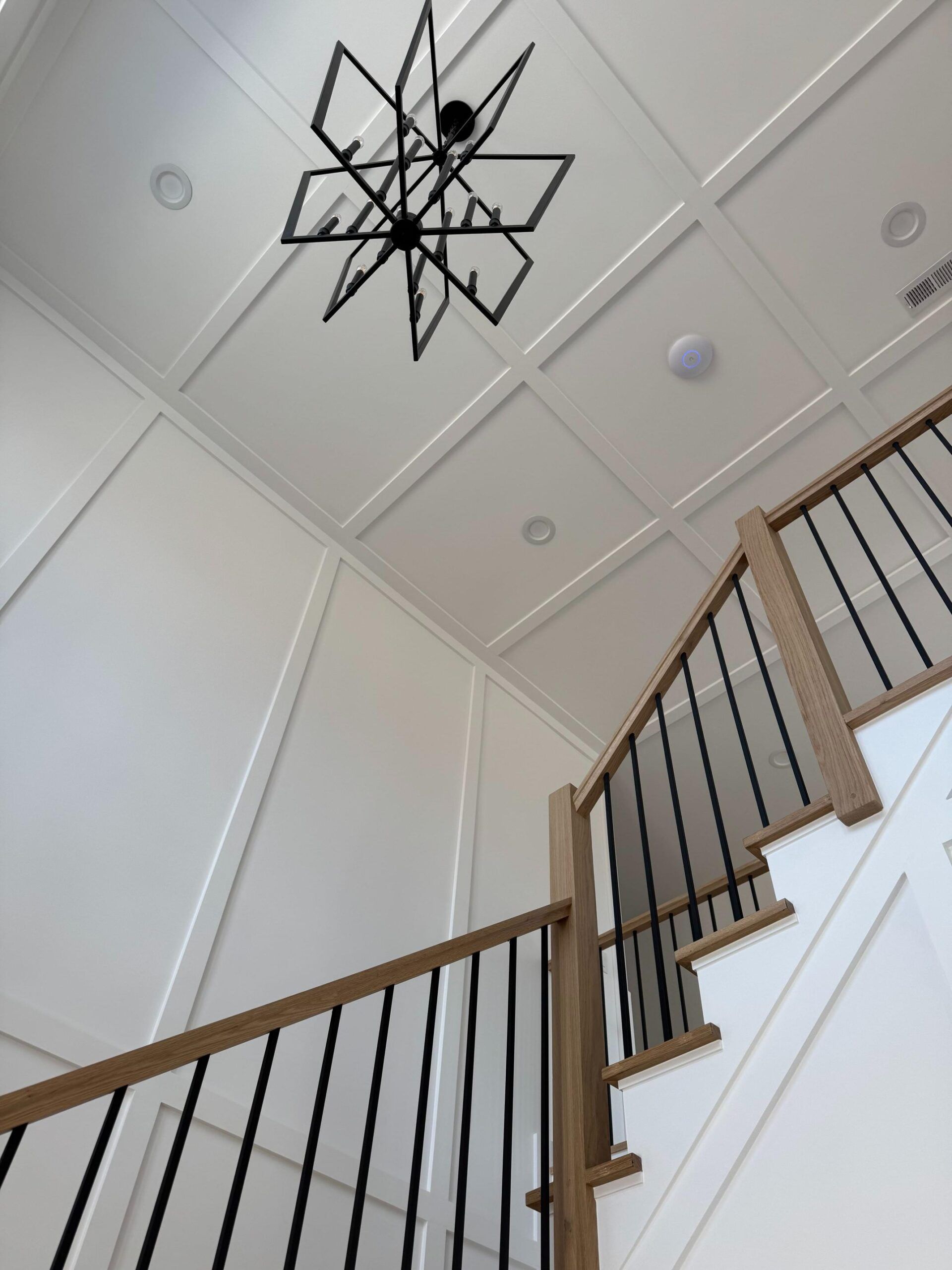 Staircase with black railing and light wooden accents; white walls and ceiling with a geometric chandelier.