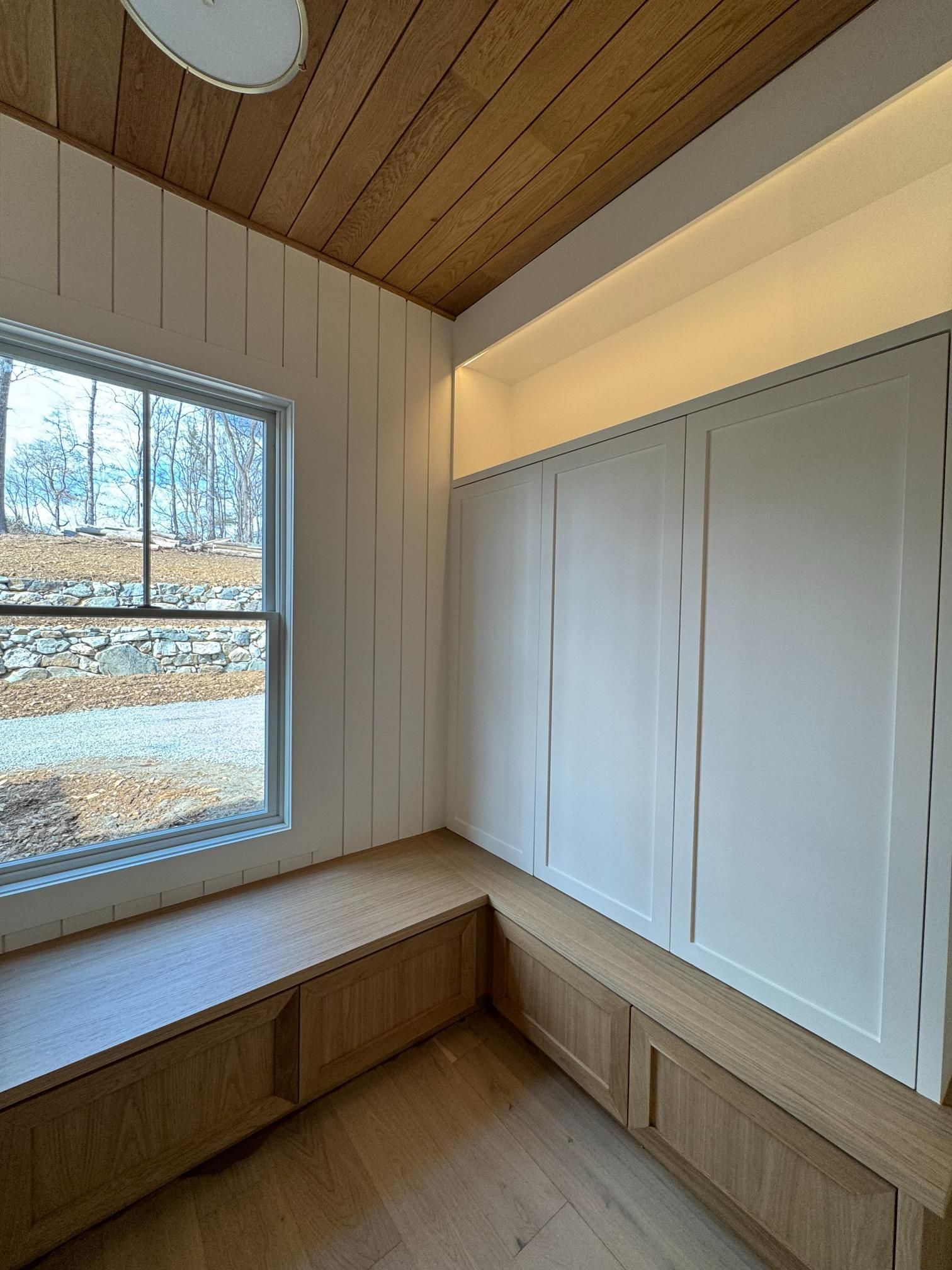 Cozy room corner with a window and a built-in wooden bench with storage. White walls, wood ceiling.
