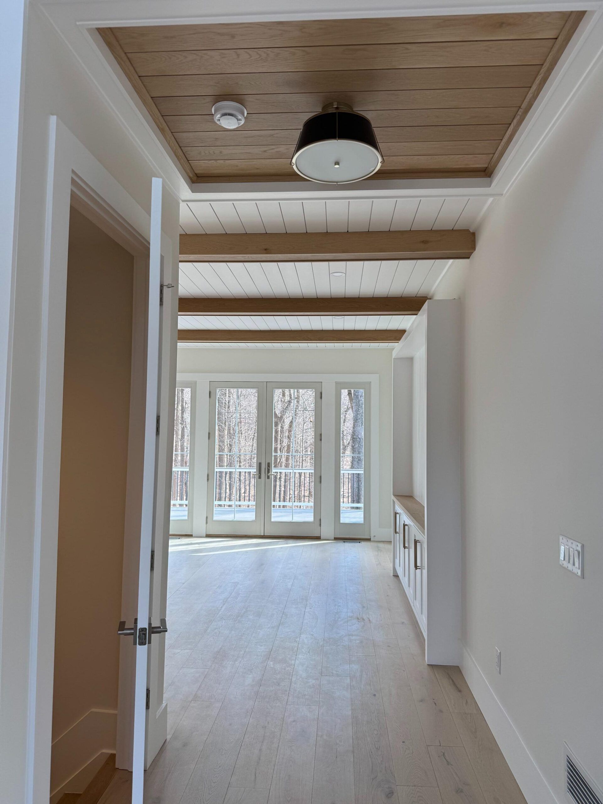 Hallway with a wood-paneled ceiling, a black light fixture, and white walls leading to a room with glass doors.
