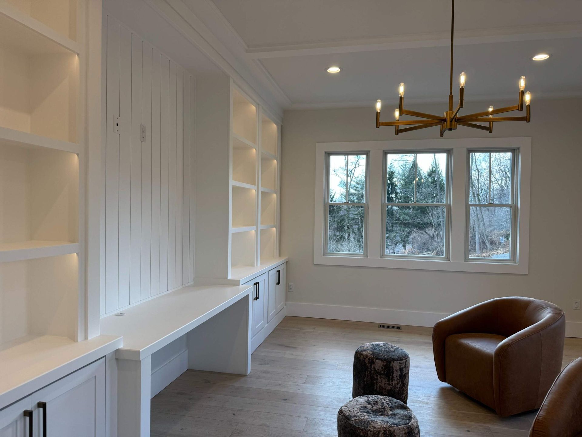 Bright, white built-in shelving and desk, facing windows, with a chandelier, and leather chair.