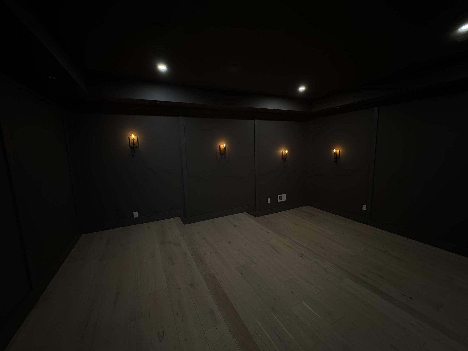 Dark home theater room with wooden floor, black walls, sconces, and recessed lights.
