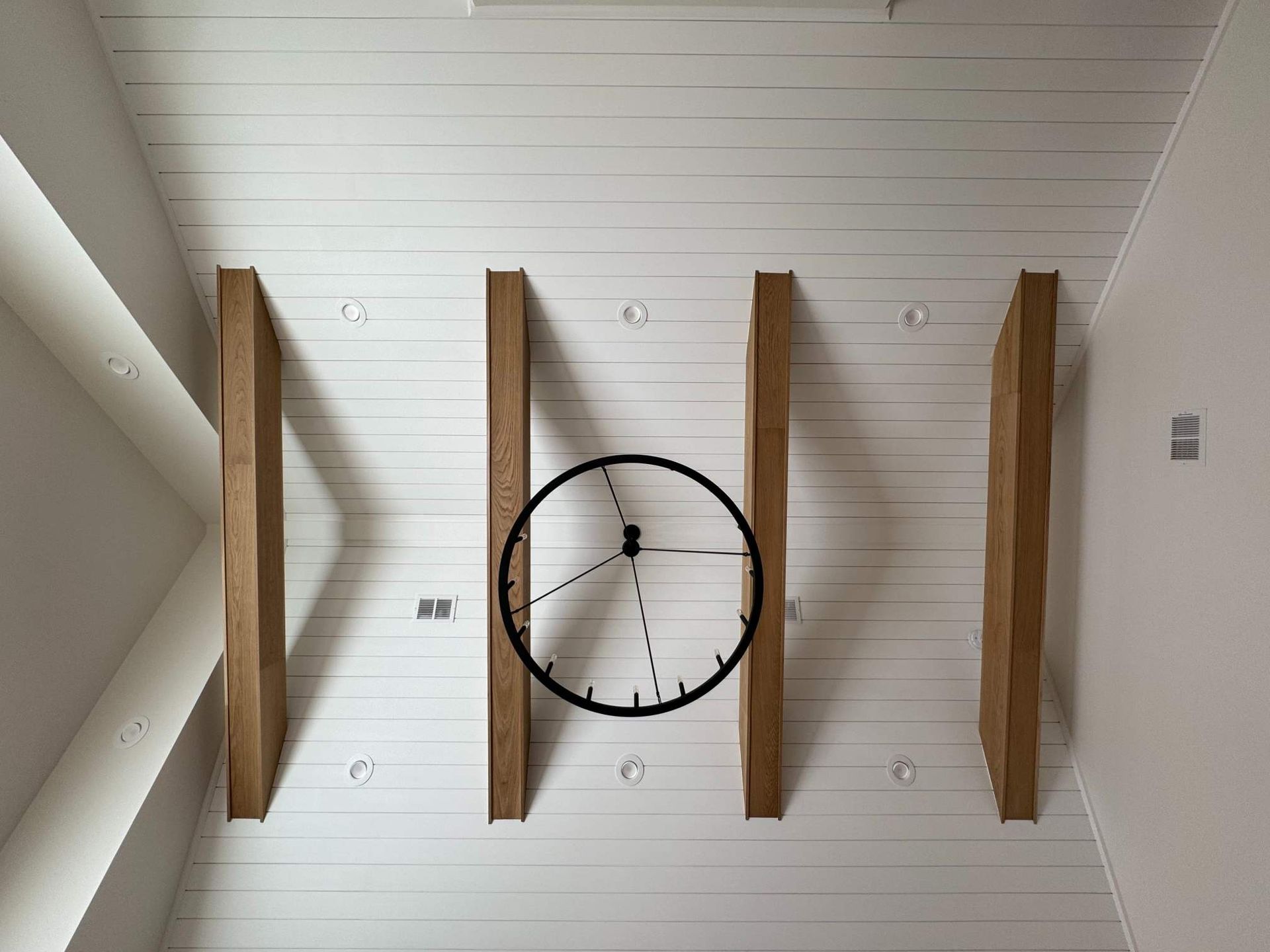Ceiling with white planks, four wood beams, a black clock, and recessed lighting.