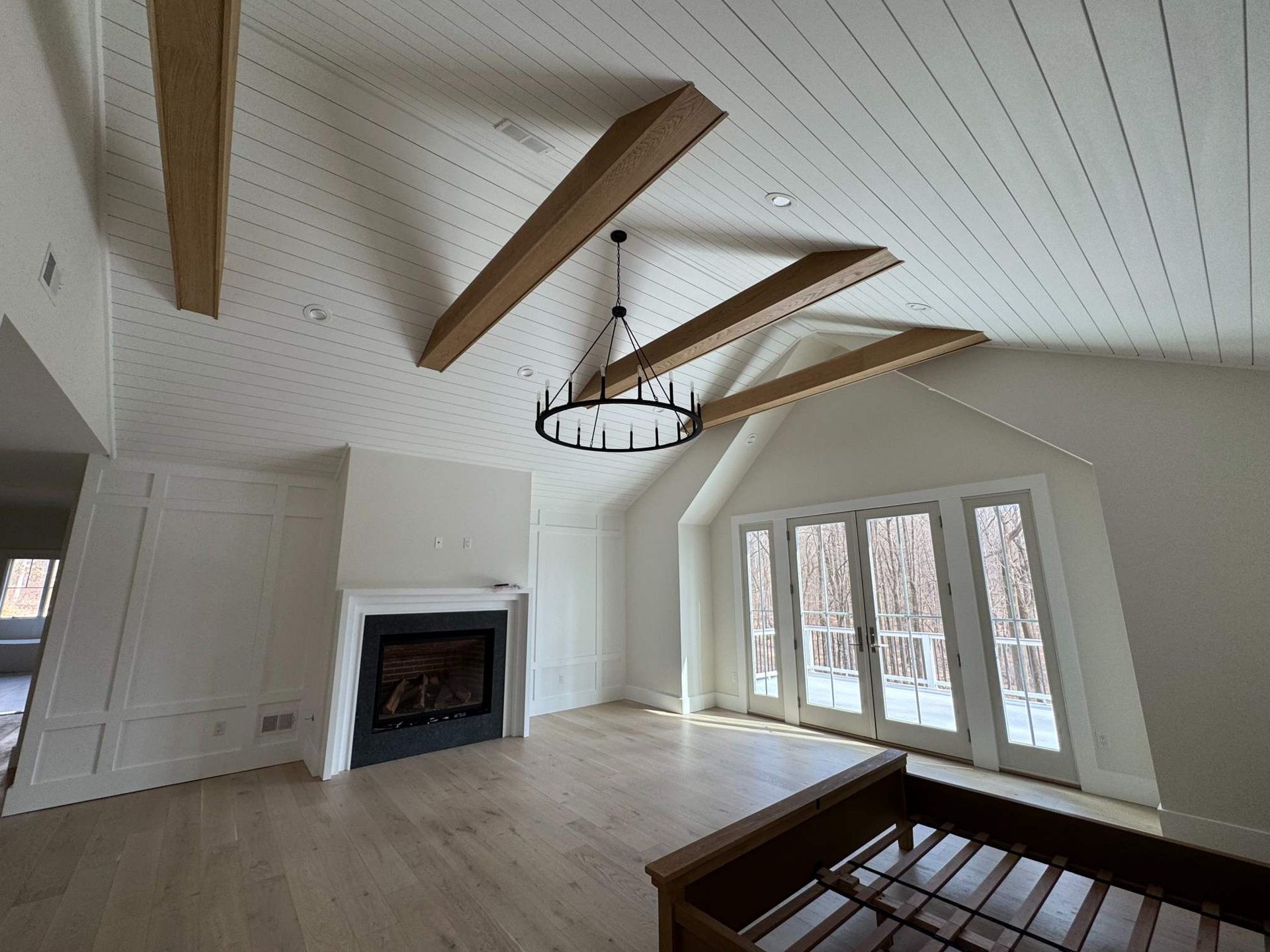Spacious white room with wooden beams, fireplace, and French doors leading to a wooded view.