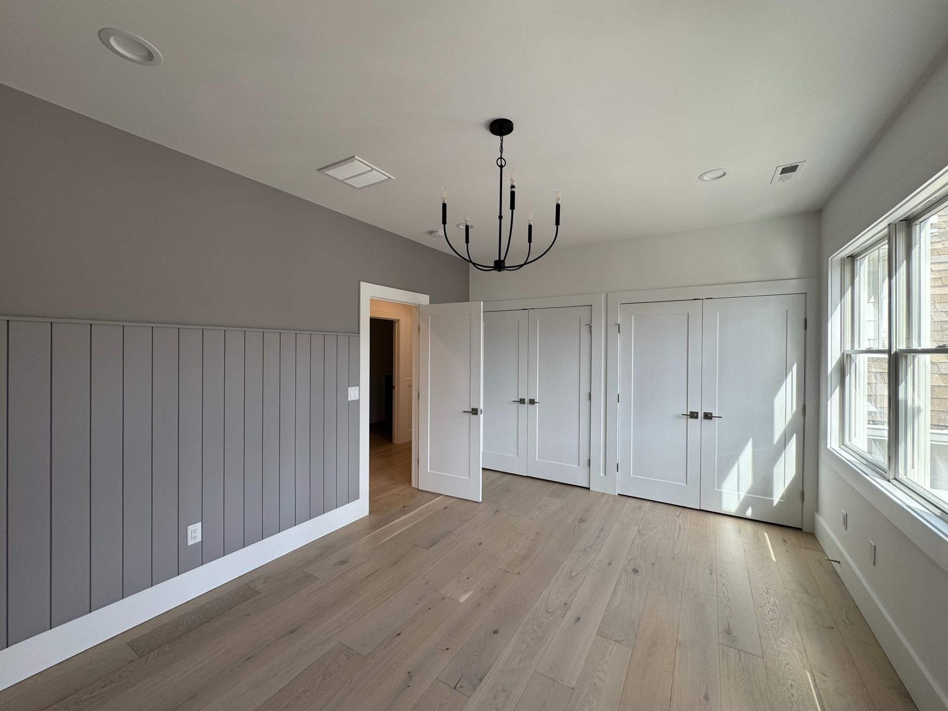 Empty bedroom with gray paneling, white doors/trim, wood floors, and black chandelier.