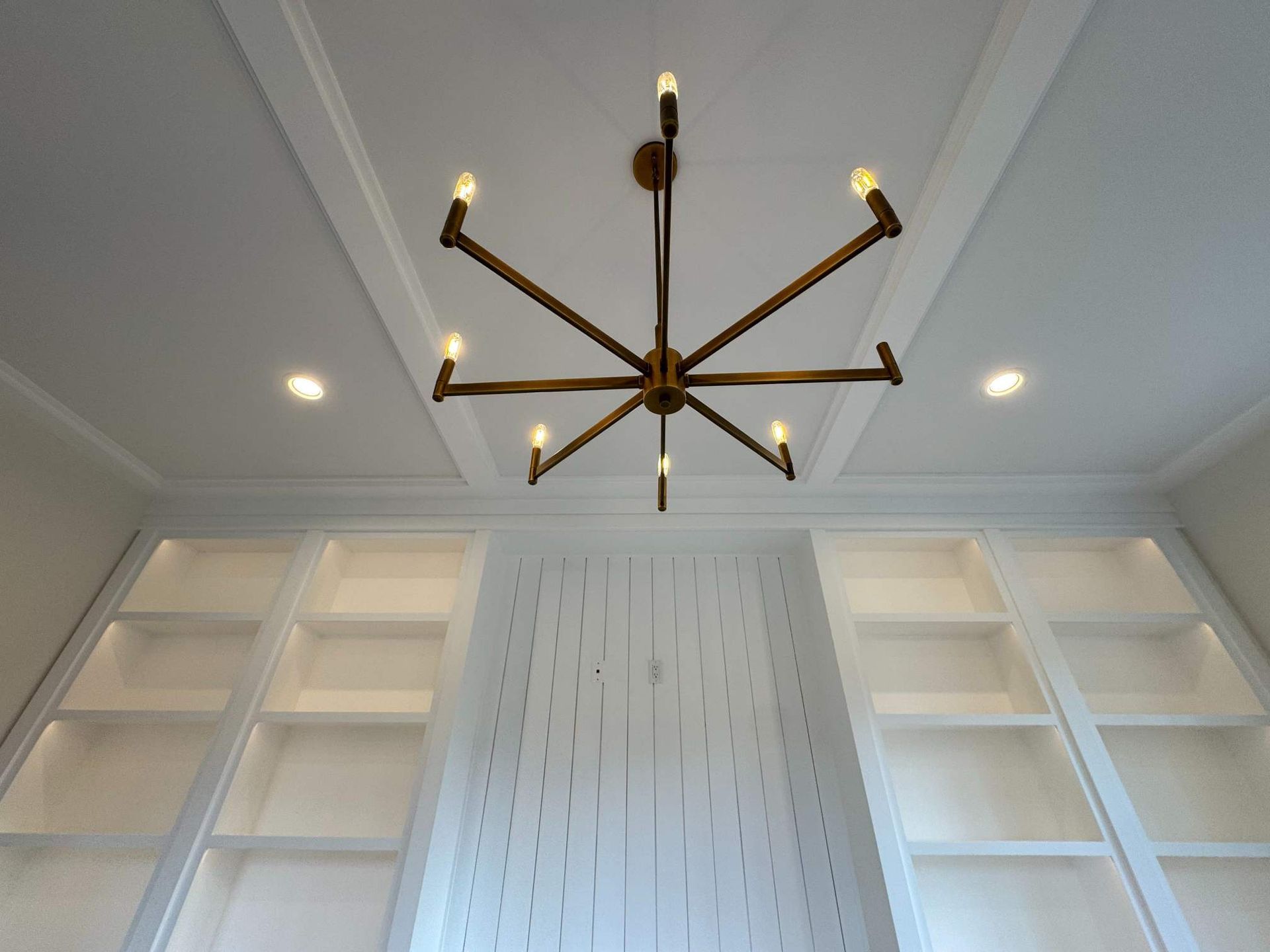 A modern, gold chandelier hangs in a room with white built-in shelves and a white paneled wall.