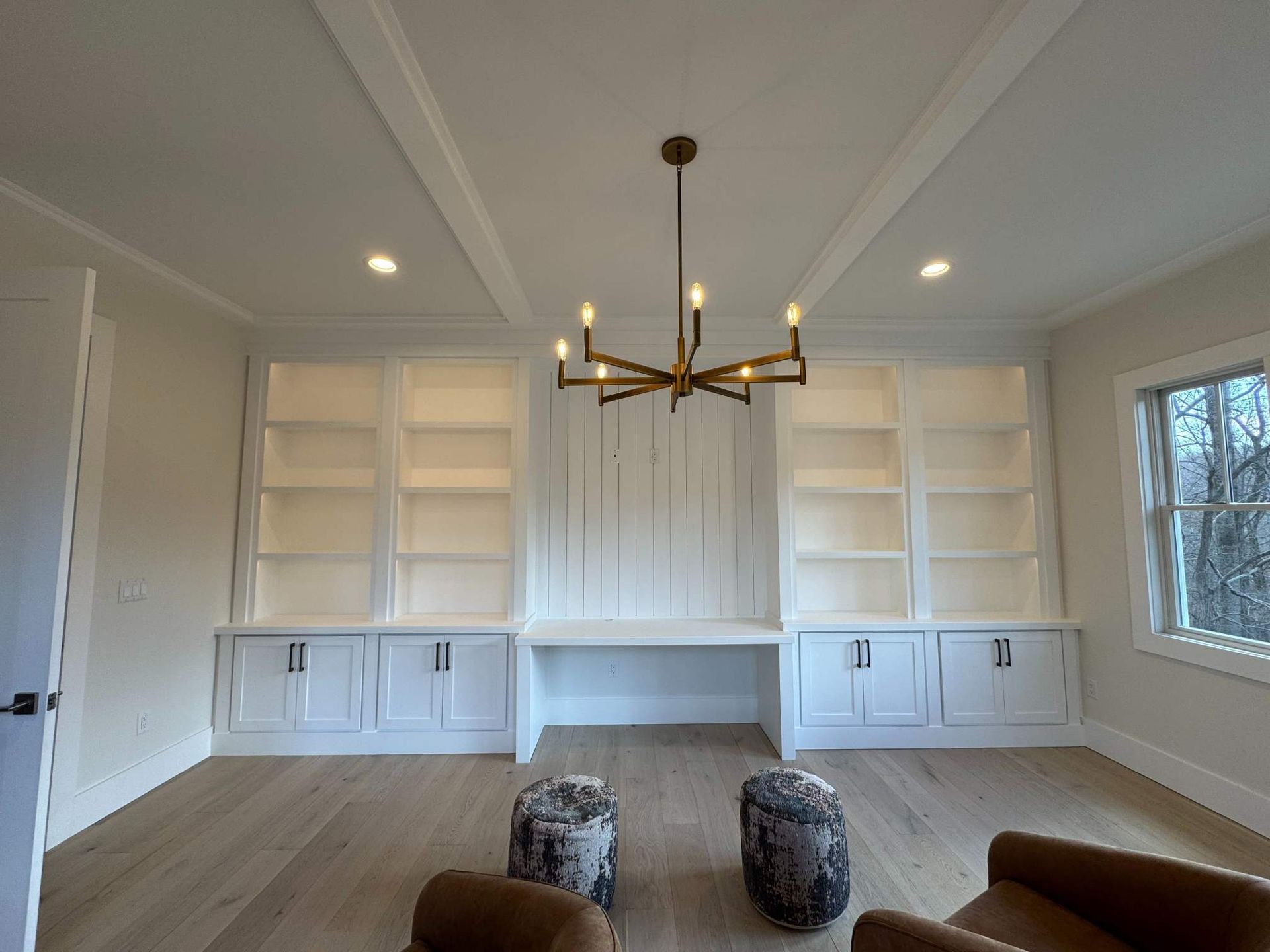 Built-in white bookshelves and desk in a room with hardwood floors, chandelier, and two patterned ottomans.