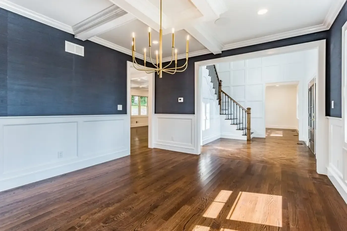 Empty dining room with dark blue walls, white trim, gold chandelier, and hardwood floors.