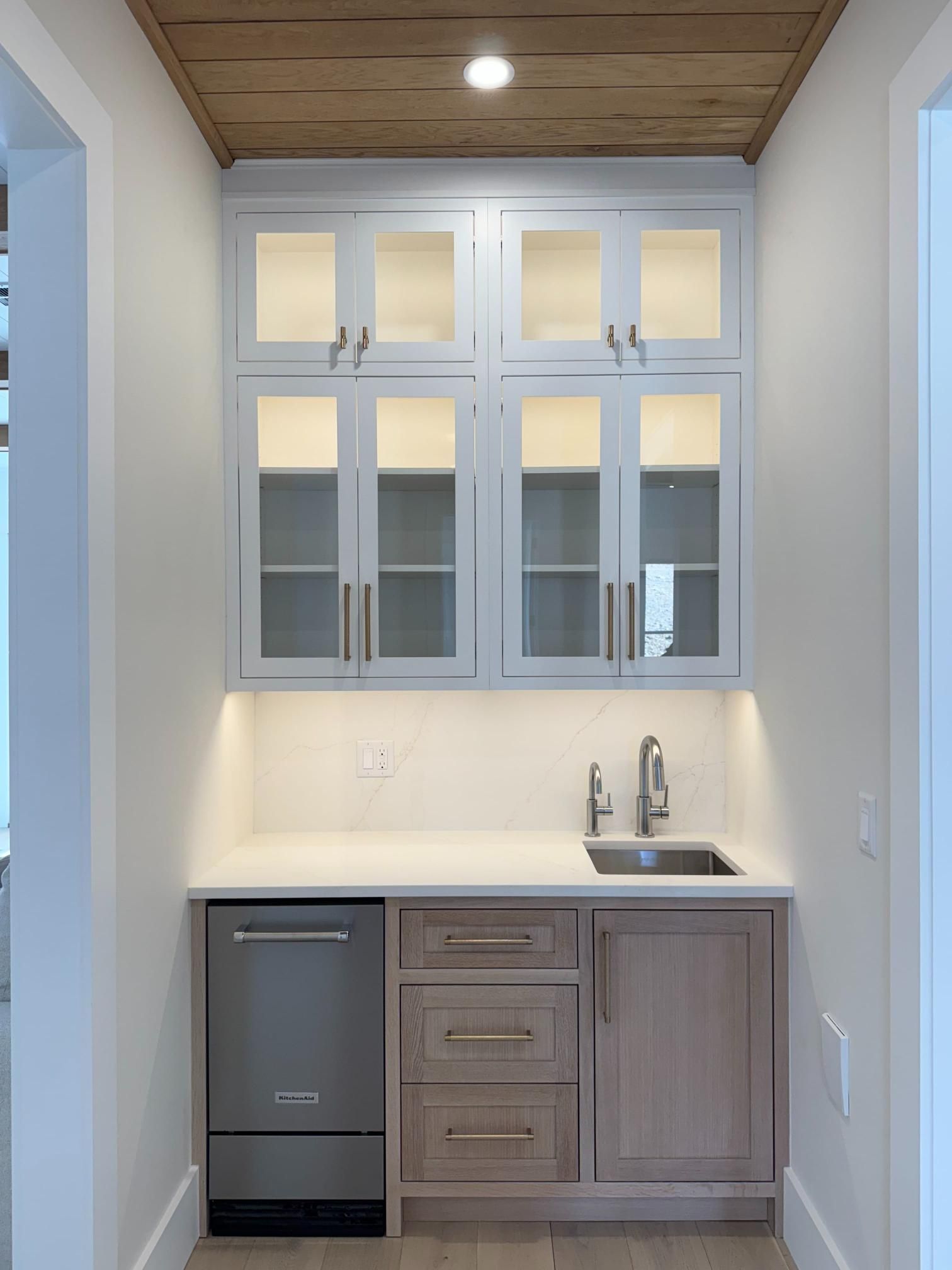 A built-in wet bar with a small sink, cabinets, and a mini-fridge in a white hallway.