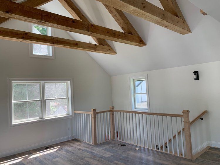 Interior with exposed wooden beams, white walls, and a staircase with a railing. Windows provide natural light.