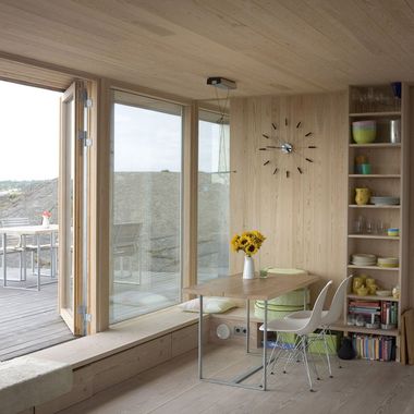 Intérieur en bois avec une table, des chaises, des étagères et une porte vitrée donnant sur une terrasse.