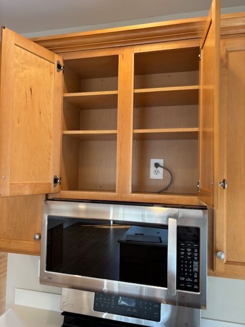 Over-the-range microwave below open light wood kitchen cabinets with interior shelves, next to a power outlet.